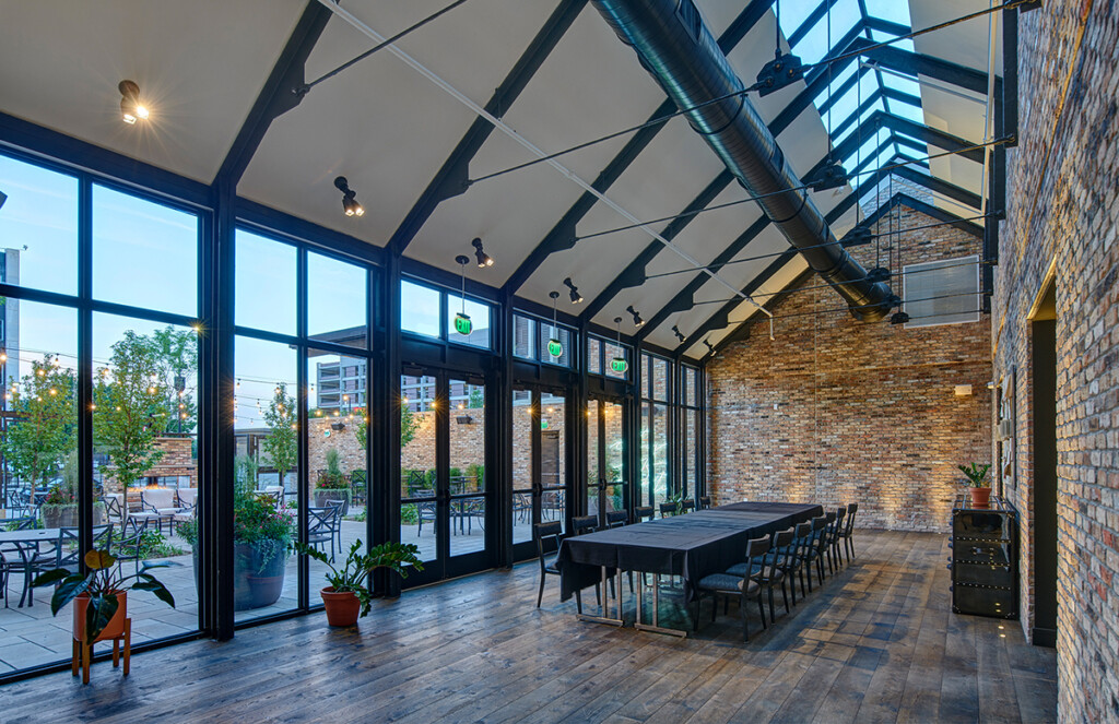 A long table sits below the atrium with angled windows on the ceiling and large doors opening to a brick patio